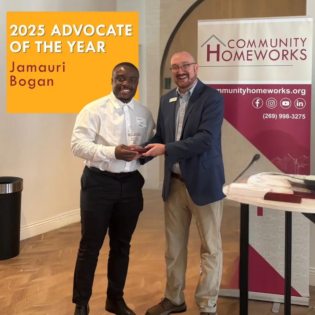 A man in a white shirt and black trousers accepts an award from a man in a blue blazer at a Community Homeworks event, with text overlay reading “2025 Advocate of the Year Jamauri Bogan.”.