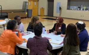 Seven people sit around a round table in a gym during a Community Homeworks Kalamazoo discussion.