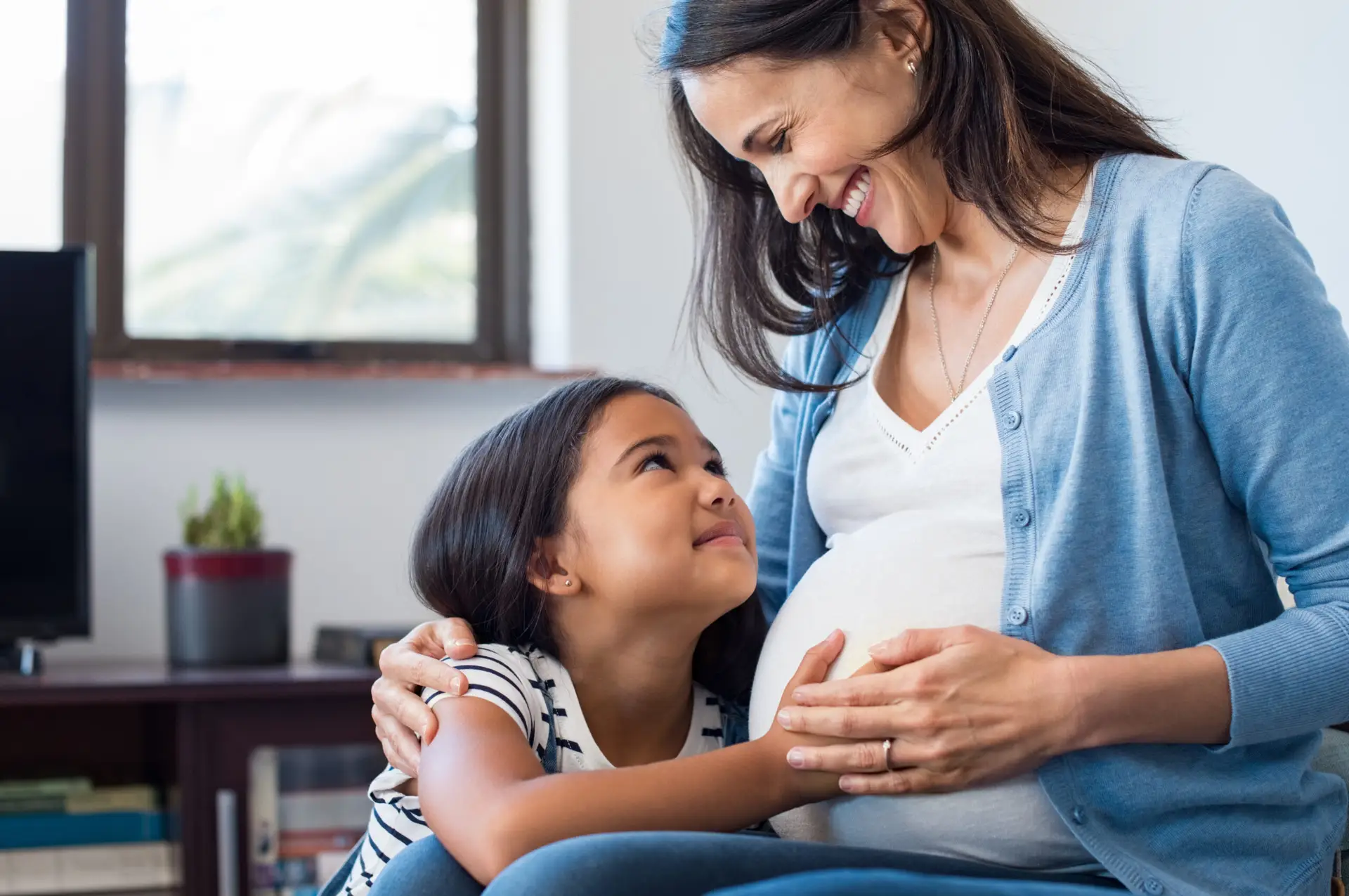 A smiling pregnant woman sits with her daughter resting her head on her stomach, a scene reflecting the heart of Community Homeworks Kalamazoo.