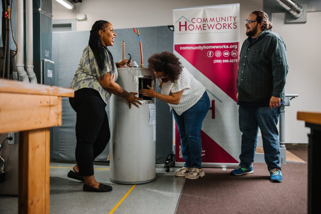 Two women inspect a water heater while a man in safety glasses watches in a Community Homeworks Kalamazoo workshop.