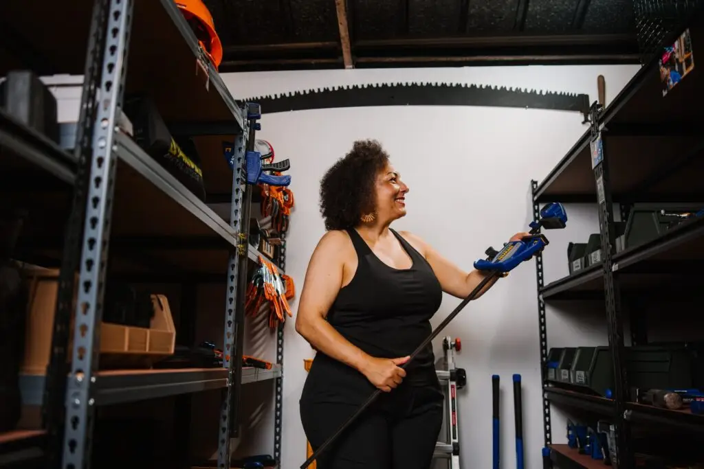 A woman smiles while holding a cordless power tool in a Community Homeworks Kalamazoo workshop with metal storage shelving.