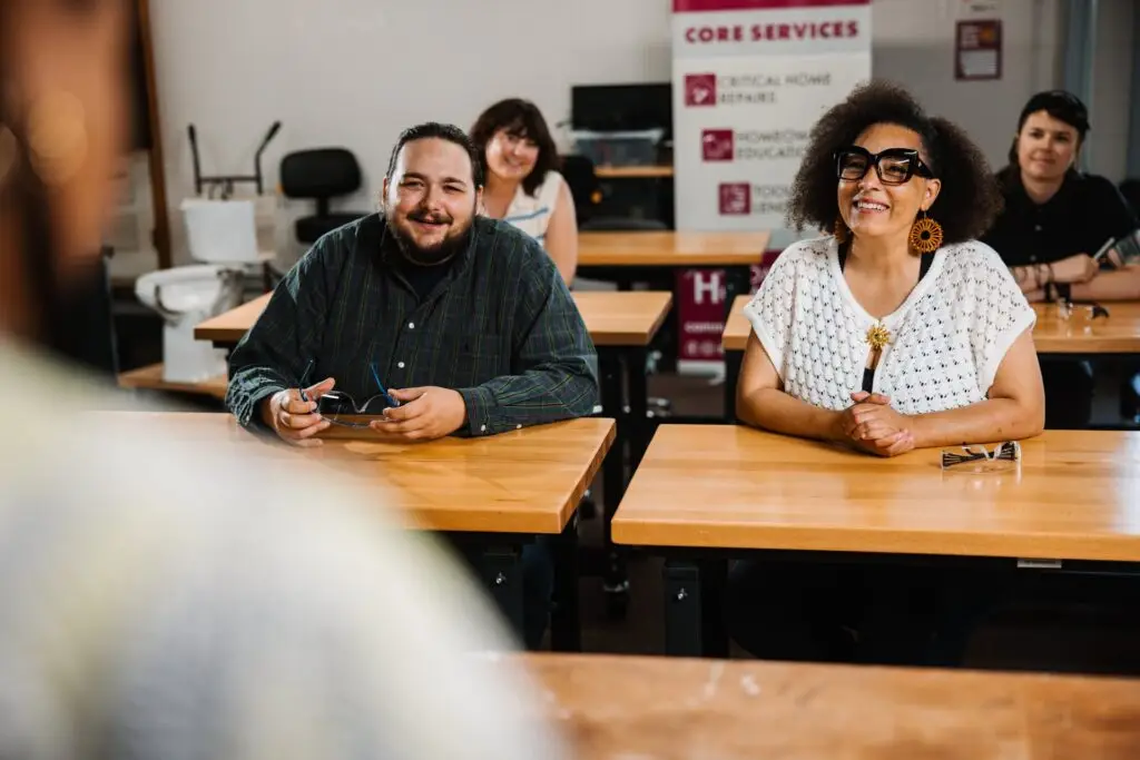 Three adults sit at wooden desks in a Community Homeworks Kalamazoo classroom, listening to a presenter.