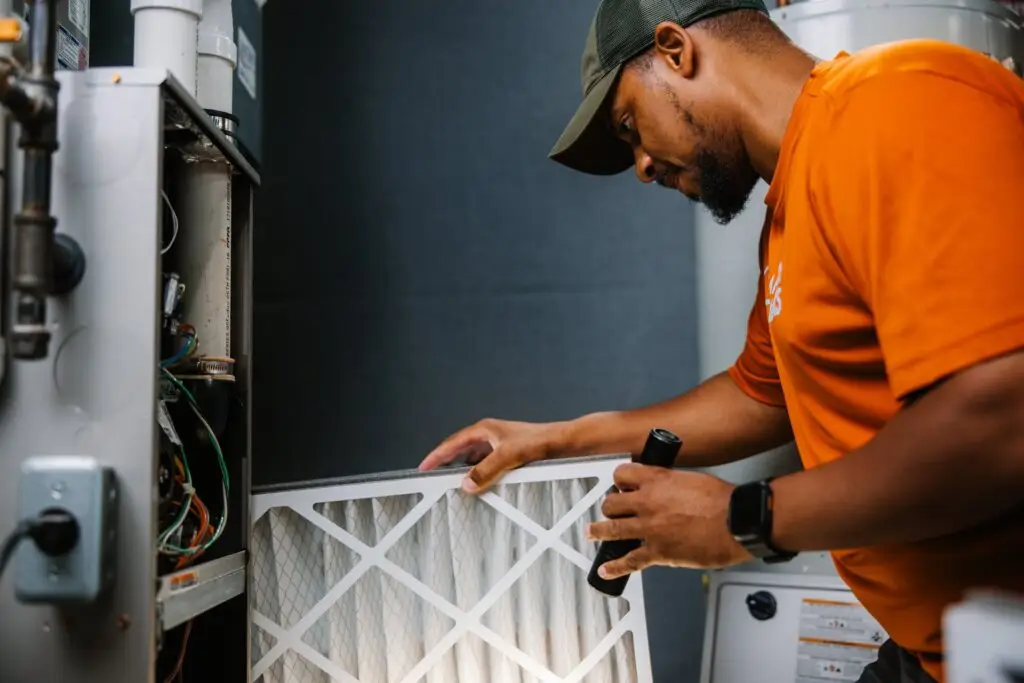 A Community Homeworks technician in an orange shirt inspects a furnace air filter with a flashlight in Kalamazoo.