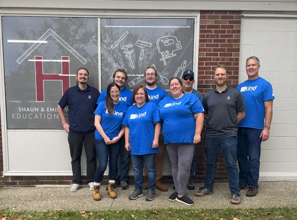 Eight people from Community Homeworks stand in a row in front of a building window, most wearing blue t-shirts with the text “kind” on the front.