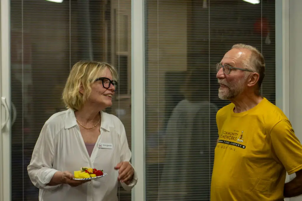 A woman holding a plate of fruit talks to a man in a yellow T-shirt at the Community Homeworks Kalamazoo office.